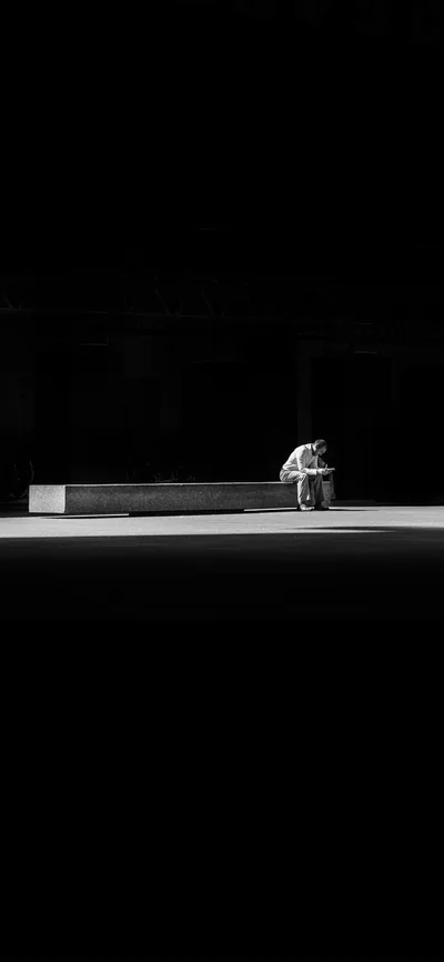 Light falls only on a strip of pavement where a man sits hunched at the end of a long concrete bench, the entire top half solid black for a clock.