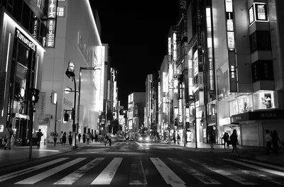 Crosswalk stripes fill the monochrome foreground as lit storefronts line both sides of a wide avenue, converging on a dark vanishing point at centre.