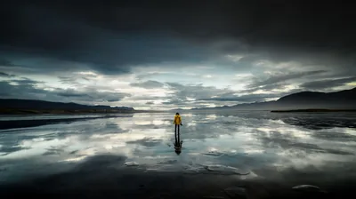 Wet tidal flats mirror a bank of grey-blue cloud around a lone yellow raincoat, low hills holding down the right end of the horizon.