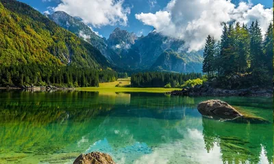 Clear green water shows every stone on the lakebed, with a single boulder mid-frame and grey Alpine summits rising behind sunlit spruce forest.