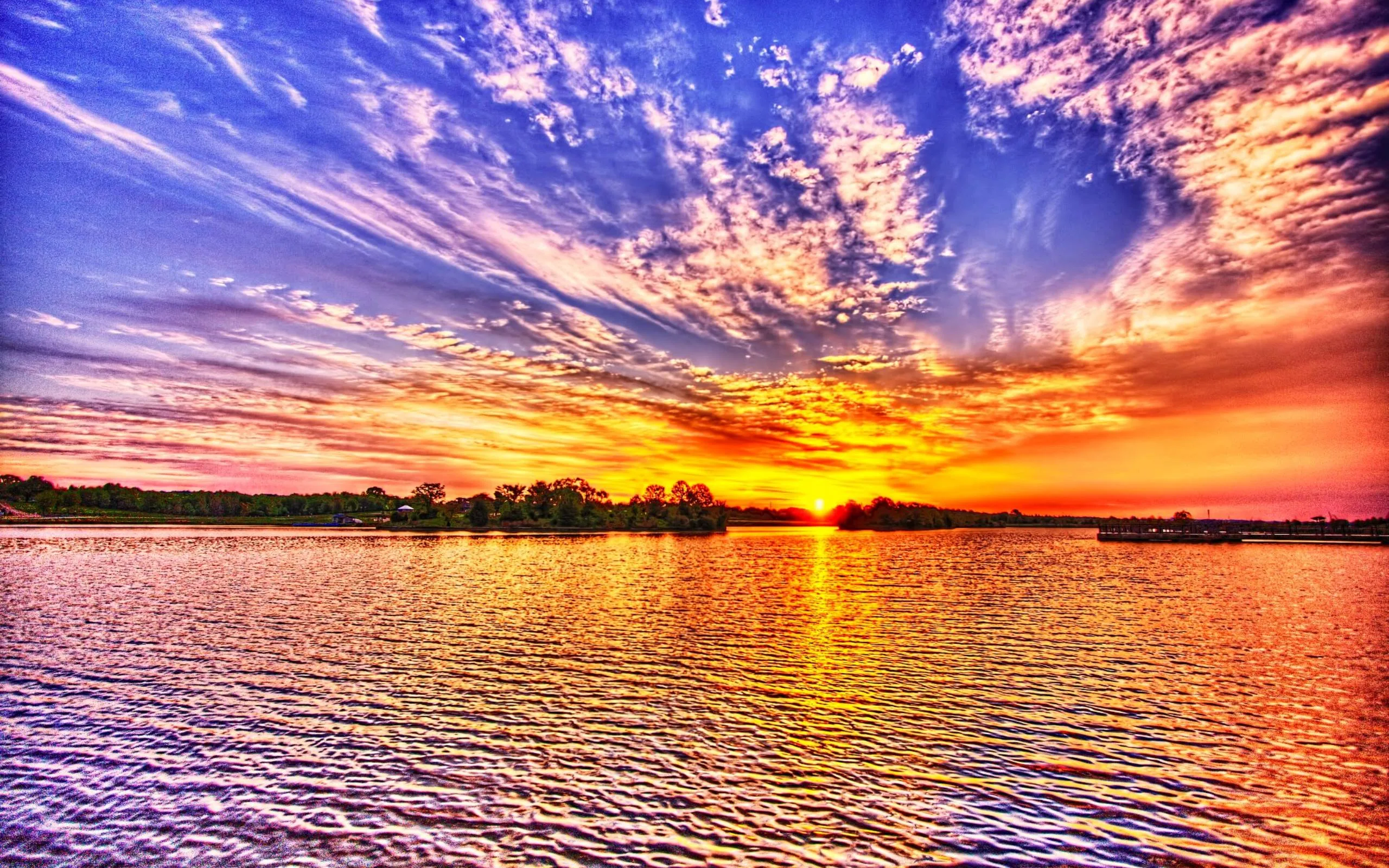 An HDR sunset saturates every cloud band in orange, pink and blue above a lake, the ripples carrying the colour down the frame.