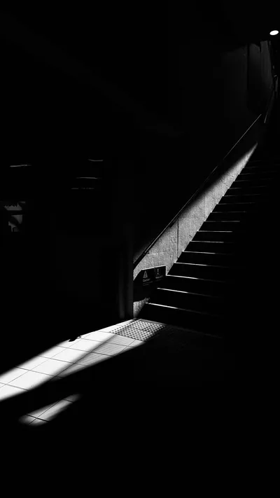 A staircase climbs into shadow on the right while window light falls in one bright patch on the floor below, the rest of the monochrome frame left black.