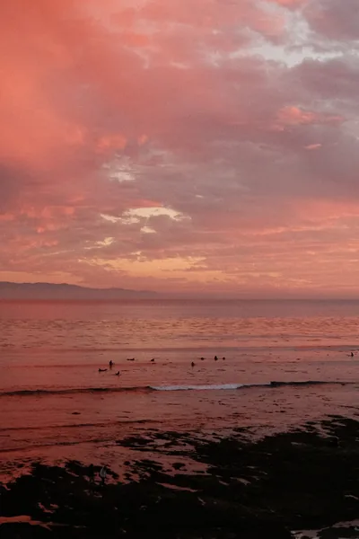 Heavy rose-coloured clouds hang over a calm bay dotted with surfers waiting beyond the shore break, dark rocks anchoring the bottom edge.