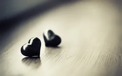 Two small glossy black hearts rest on pale wood grain, the nearer one sharp and the second blurred into warm bokeh behind it.