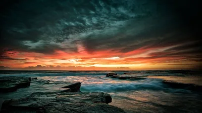 Waves wash over a shelf of dark rock while a heavy cloud bank burns orange and red along the horizon.