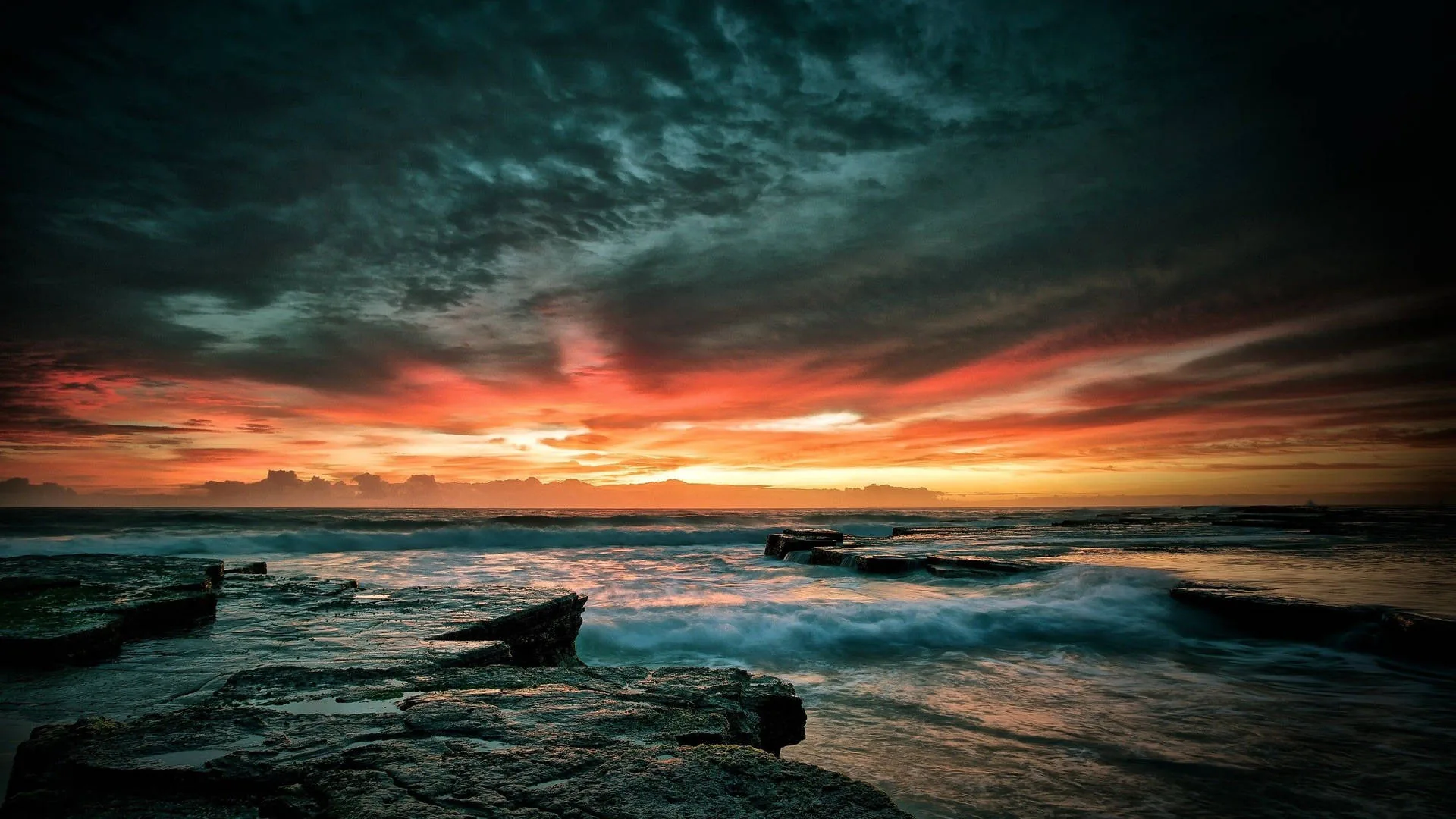 Waves wash over a shelf of dark rock while a heavy cloud bank burns orange and red along the horizon.