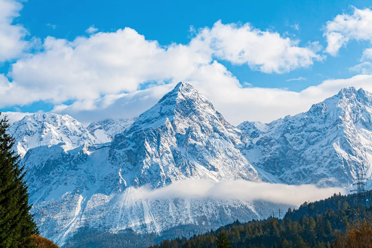A band of white cloud wraps the base of a snow-covered massif, with dark pines along the bottom edge and blue sky breaking through above.