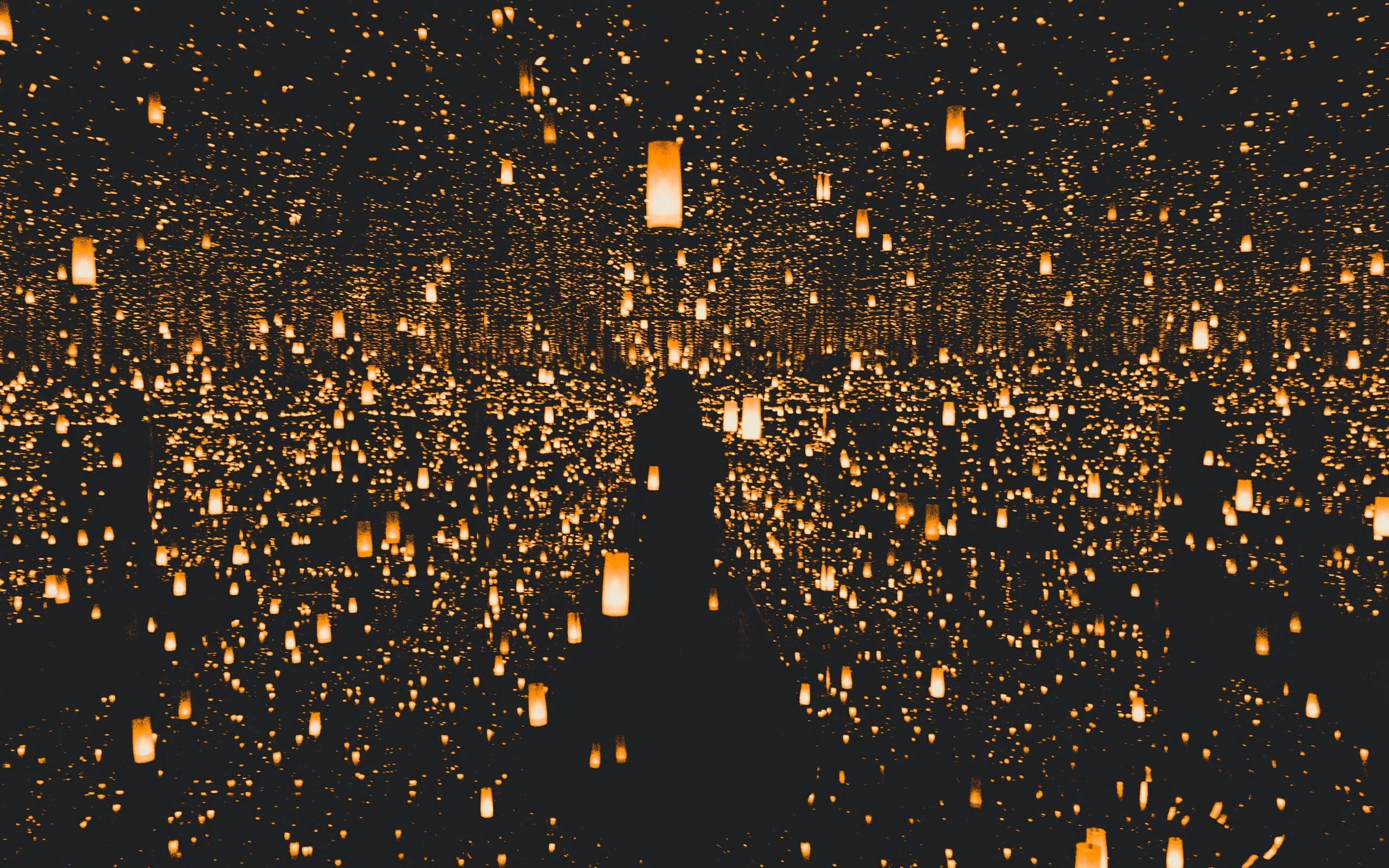 Hundreds of warm paper lanterns hang and repeat into mirrored darkness, a lone silhouette standing among them at the centre.