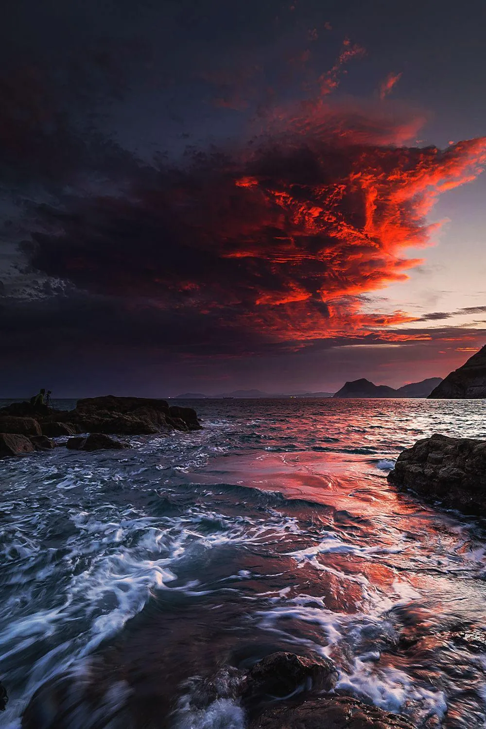 A long-exposure coast at dusk where blurred water threads between dark rocks and one crimson cloud burns over the headland.