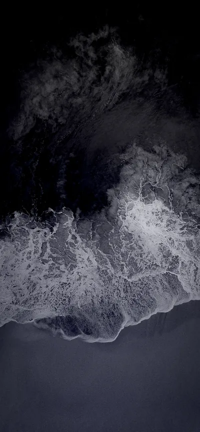 An aerial strip of coastline where white foam curls into the ink-dark water, the sand below reading as smooth cool grey.