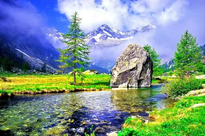 A house-sized rock splits the stream below snow peaks wrapped in cloud, pebbles showing through shallow water in this heavily saturated postcard-style scene.