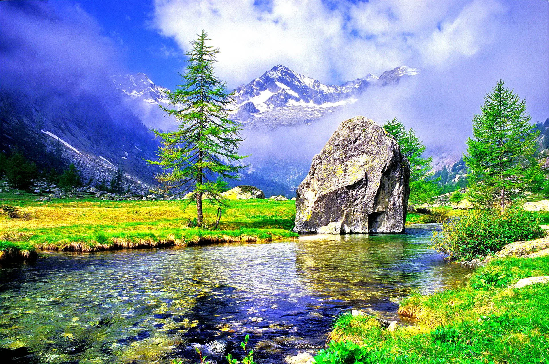 A house-sized rock splits the stream below snow peaks wrapped in cloud, pebbles showing through shallow water in this heavily saturated postcard-style scene.