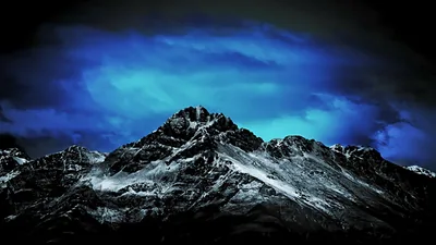 A jagged snow-streaked summit rises against heavy blue cloud, the foreground slopes falling away into black.