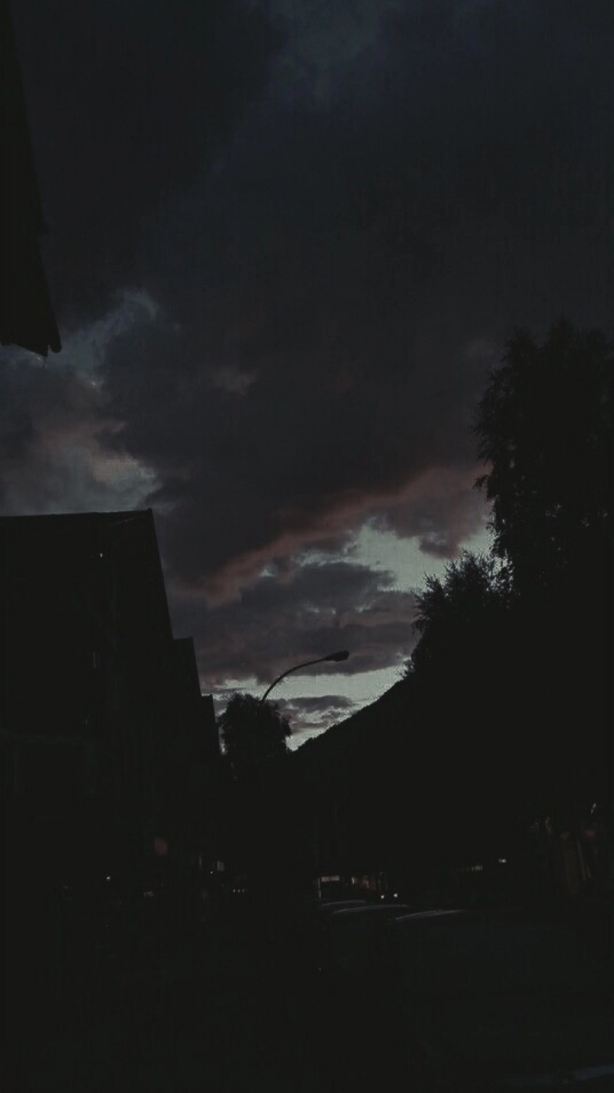 The last purple light breaks through heavy cloud over a street, rooftops and trees already fallen into silhouette.