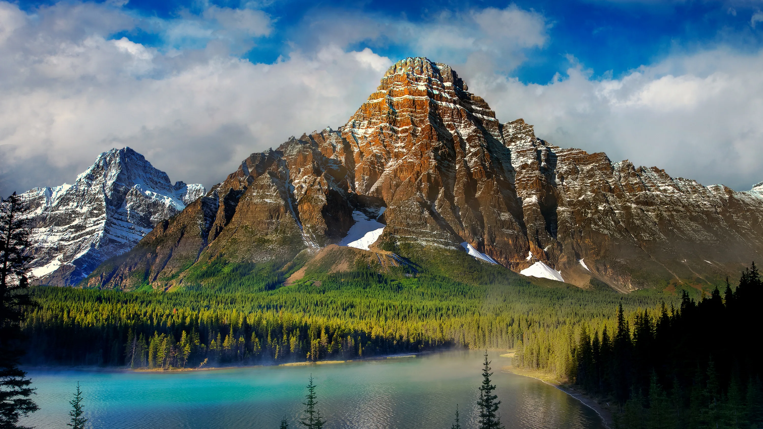 A banded rock pyramid towers over pale blue water and unbroken forest, cloud shadow keeping the near shore in cool dark tones.