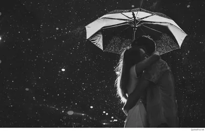 Monochrome shot of two people kissing beneath an open umbrella while snowflakes drift through the dark, the flakes catching the flash as bright specks.