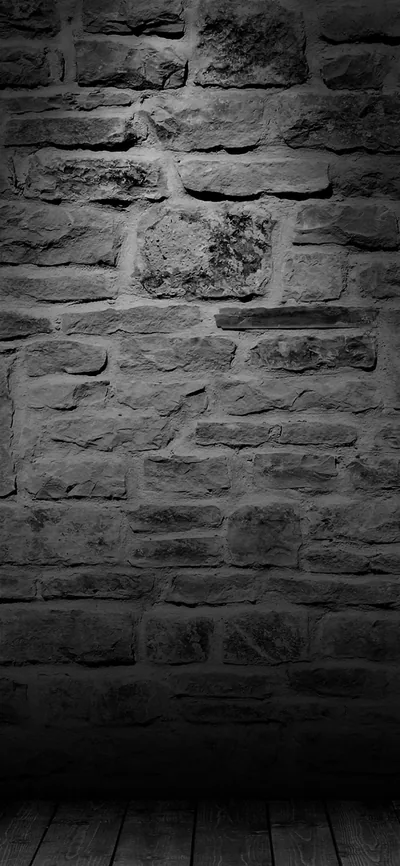 A rough old stone wall in grey monochrome meets a plank floor at the bottom of the frame, light falling from above.