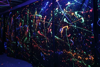 Fluorescent paint splatters in green, pink and orange glow across black panels receding to the right, a strip of bare floor showing in the lower-left corner.