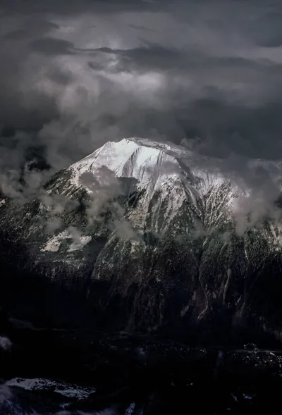 A snowy summit breaks through heavy cloud, the white ridge and its wind-blown edges the only bright thing in an otherwise slate-dark frame.