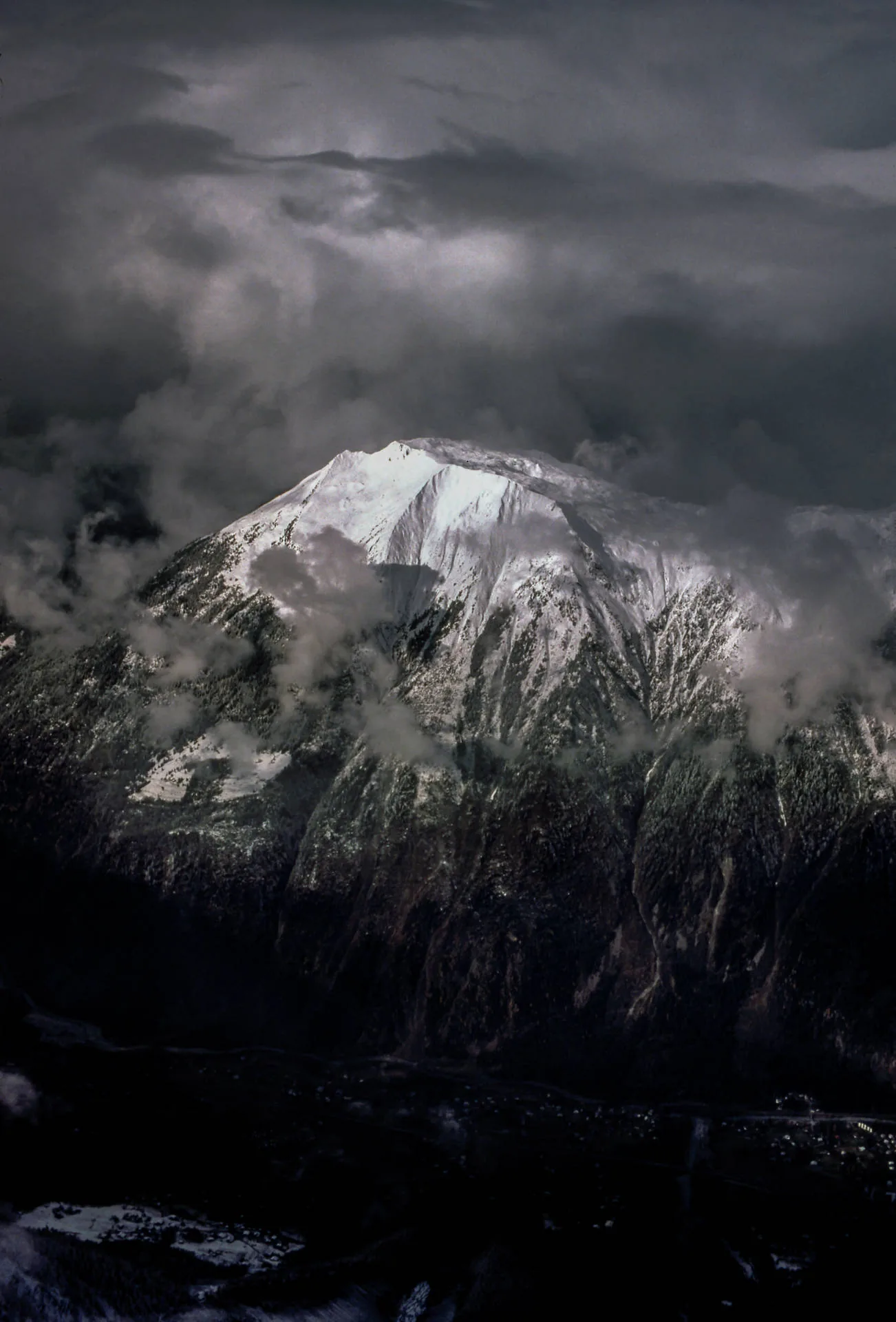 A snowy summit breaks through heavy cloud, the white ridge and its wind-blown edges the only bright thing in an otherwise slate-dark frame.