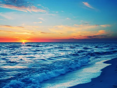 The sun touches the horizon in a band of red and orange while blue-toned waves wash up the sand, foam tracing the bottom edge.