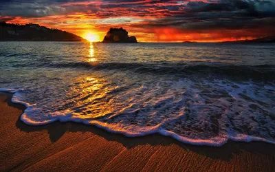 White foam curls onto wet sand while the sun drops behind a rocky islet, the whole sky banded in red and orange.