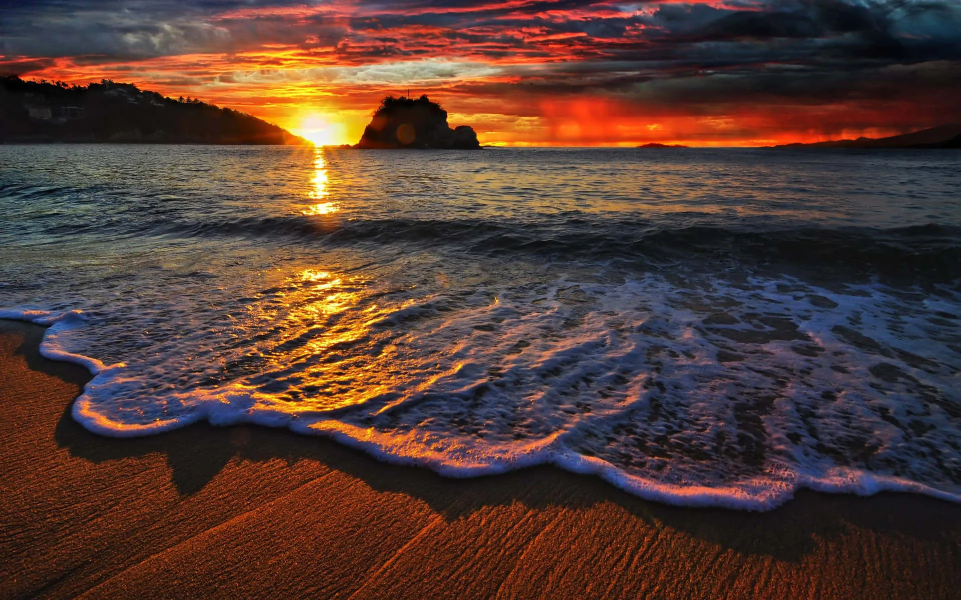 White foam curls onto wet sand while the sun drops behind a rocky islet, the whole sky banded in red and orange.
