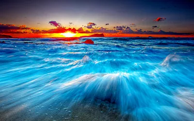 Long exposure turns receding surf into electric blue streamers over the sand while the sky burns red and orange at the horizon.