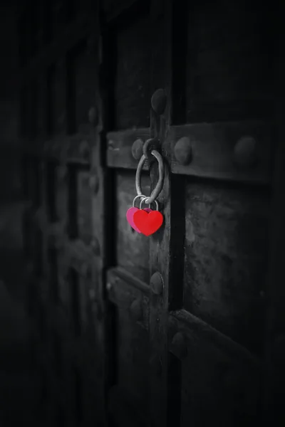 A red heart-shaped lock hangs from an iron ring on a studded black door, the only colour in a desaturated frame.
