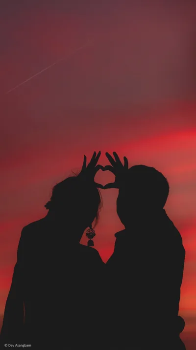 Two figures lean together and join their fingers into a heart shape, cut as pure black silhouettes against a deep red evening sky.