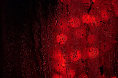 Water beads cling to a dark window while red lights blur into round bokeh spots behind it, the left edge left almost black.