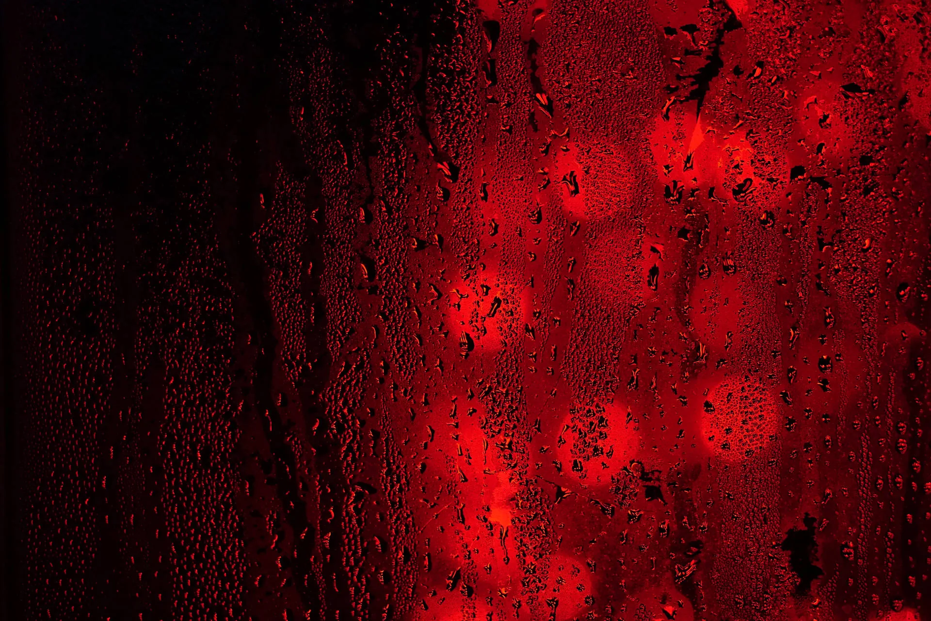 Water beads cling to a dark window while red lights blur into round bokeh spots behind it, the left edge left almost black.