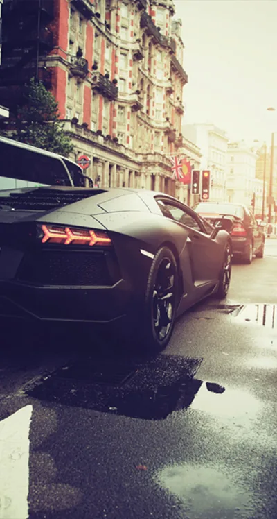 Seen from behind, a matte black Lamborghini sits kerbside on a rain-slicked street with old apartment blocks rising into hazy daylight.