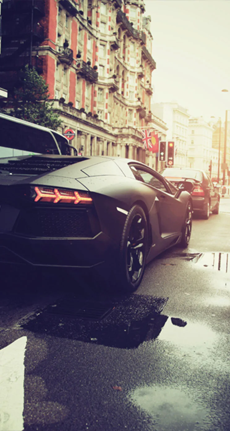 Seen from behind, a matte black Lamborghini sits kerbside on a rain-slicked street with old apartment blocks rising into hazy daylight.