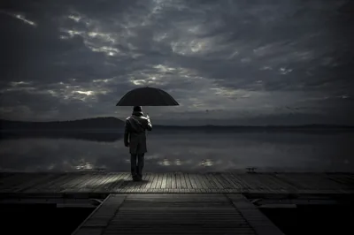 Under a heavy slate sky, someone holds a wide black umbrella at the end of a lakeside dock; the clouds break faintly in the upper left only.