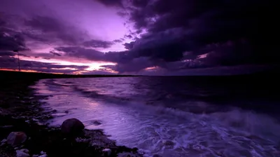 Heavy cloud turns violet over a rocky shoreline, surf breaking pale across the stones in the foreground.