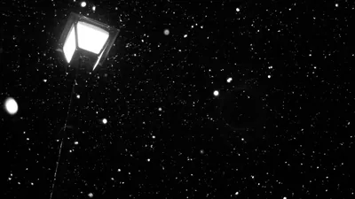 A single square street lamp glows in the top left corner while snowflakes catch the light against a completely black winter sky.