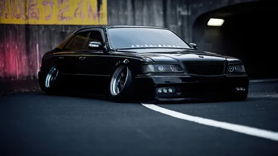 A lowered JDM saloon sits on stretched tyres in a concrete underpass, pink and yellow graffiti smeared across the wall behind it and a white lane line running past the front bumper.
