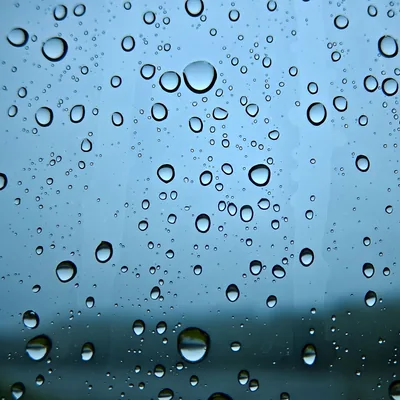 Round raindrops stick to glass in front of a pale powder-blue sky, with a soft dark band of ground blurring across the bottom quarter.