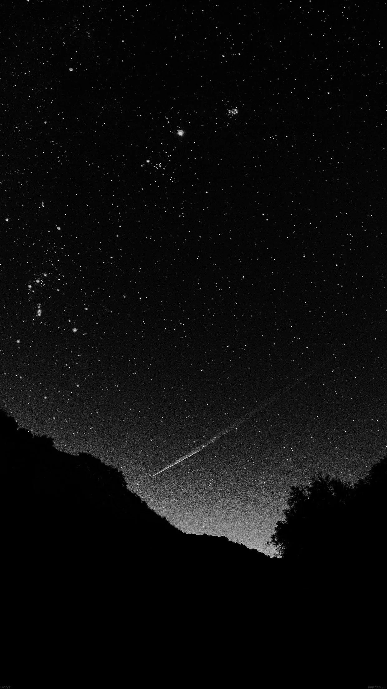 Monochrome portrait of a single meteor cutting low across the frame, a pale glow rising behind the mountain slope beneath scattered stars.