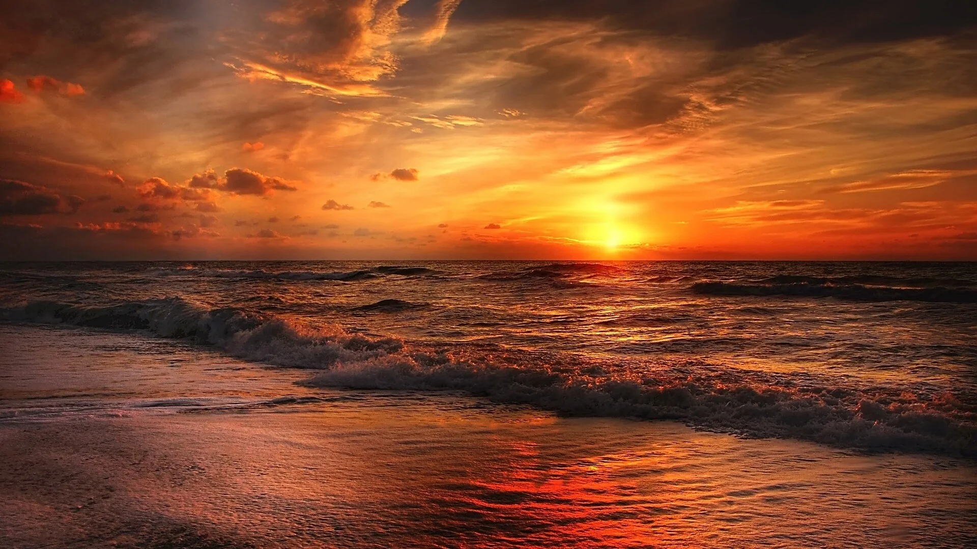 Ragged clouds smoulder in red and gold above lines of breaking waves, the light pooling in reflections on the dark wet beach.