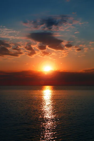 The sun burns through a gap in slate-grey clouds and drops a narrow orange path down the sea toward the bottom of the frame.