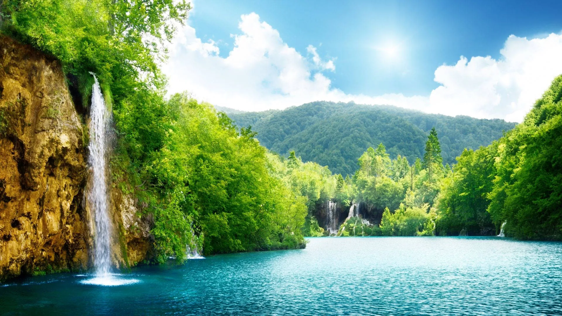 Two slender falls pour off the left cliff into blue-green water, the sun burning high at centre; midday light keeps shadows short across the forest.