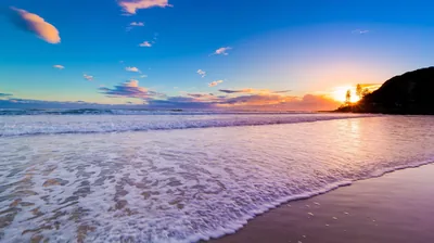 White foam fills the foreground as the sun bursts from behind a dark headland on the right, the sky above still a clean daytime blue.
