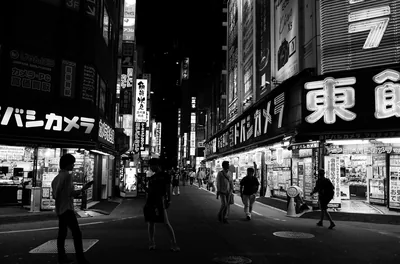 Pedestrians drift past glowing vertical shop signs on a night street, the brightest lettering stacked along the right while the road ahead sinks to black.