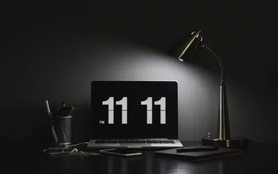 A lamp throws one cone of light across a dark desk, the laptop showing a large flip clock reading 11:11 beside a pen pot.
