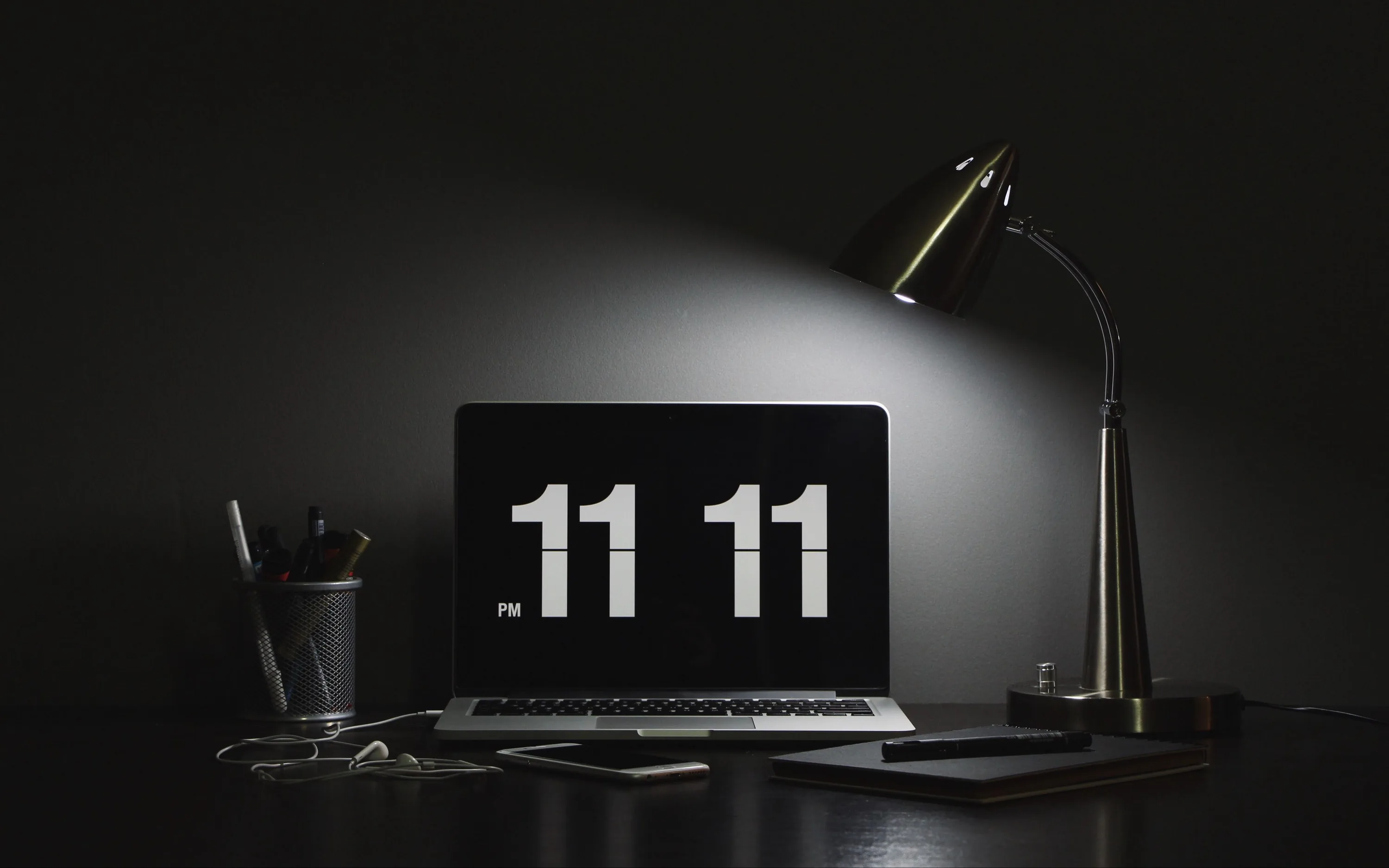 A lamp throws one cone of light across a dark desk, the laptop showing a large flip clock reading 11:11 beside a pen pot.