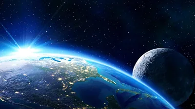 A sunrise flare breaks at the left while city lights trace the night side below, the Moon holding the right edge under a black star field.