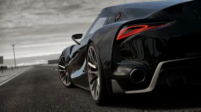 Tight crop on the rear haunch of a black coupe parked on open tarmac, a hooked red lamp glowing above the split-spoke wheel under a flat grey sky.