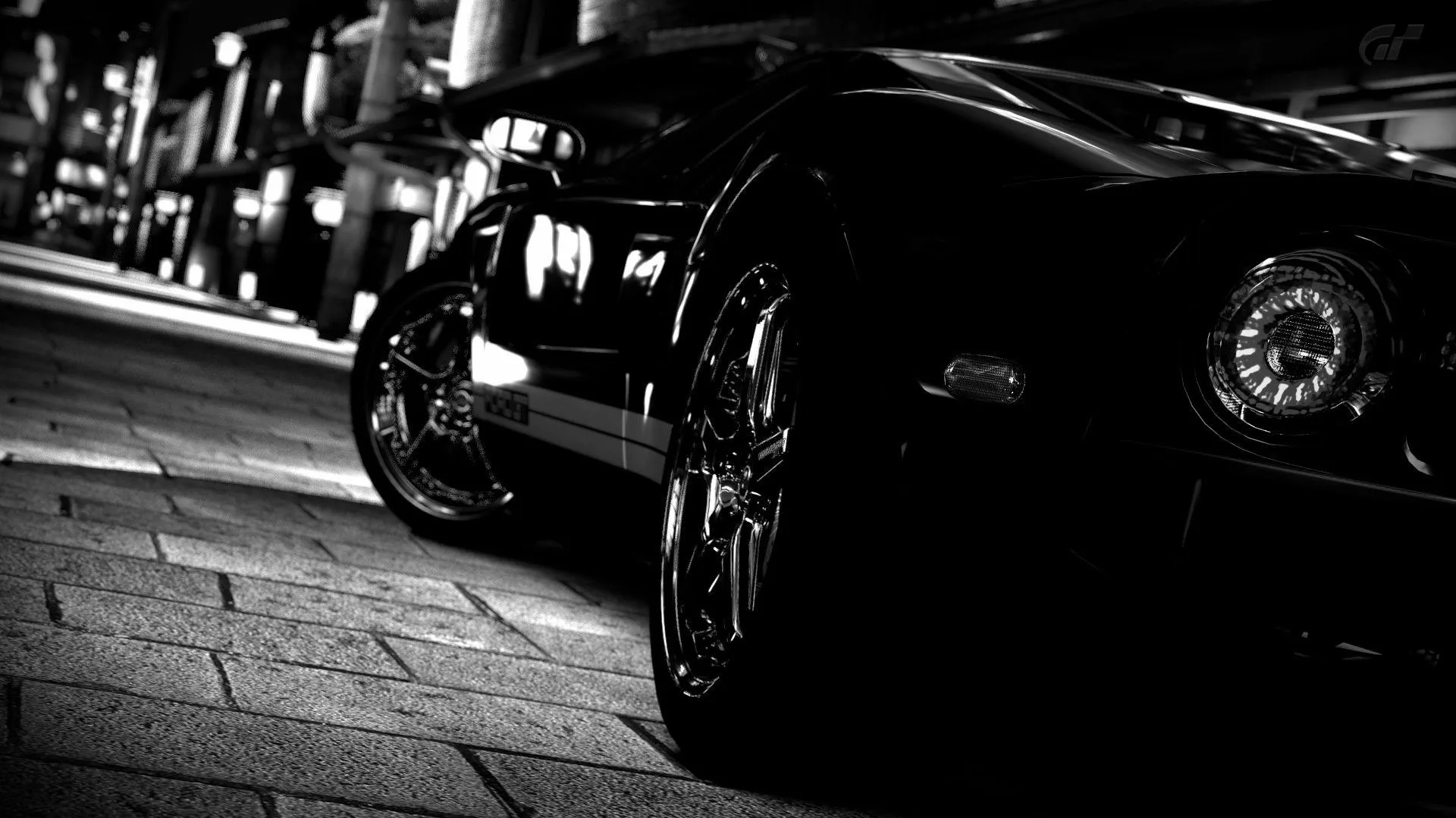 Shot from ground level behind a black supercar, the rear tyre and polished rim dominating the frame while blurred streetlights run down the pavement in monochrome.